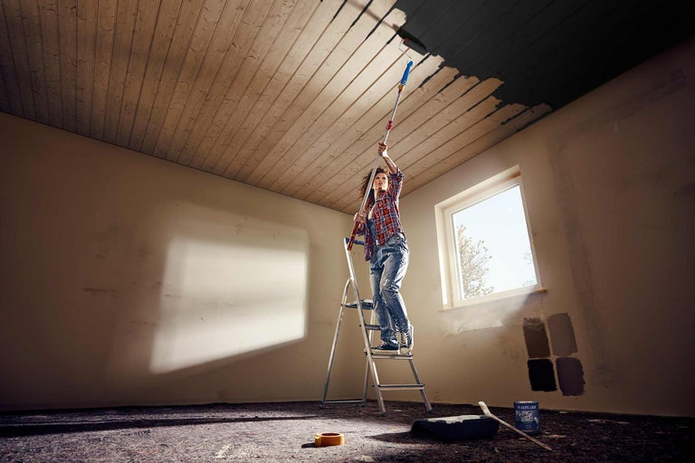 Une femme se tient sur une échelle et peint un plafond en bois avec un rouleau à peinture.