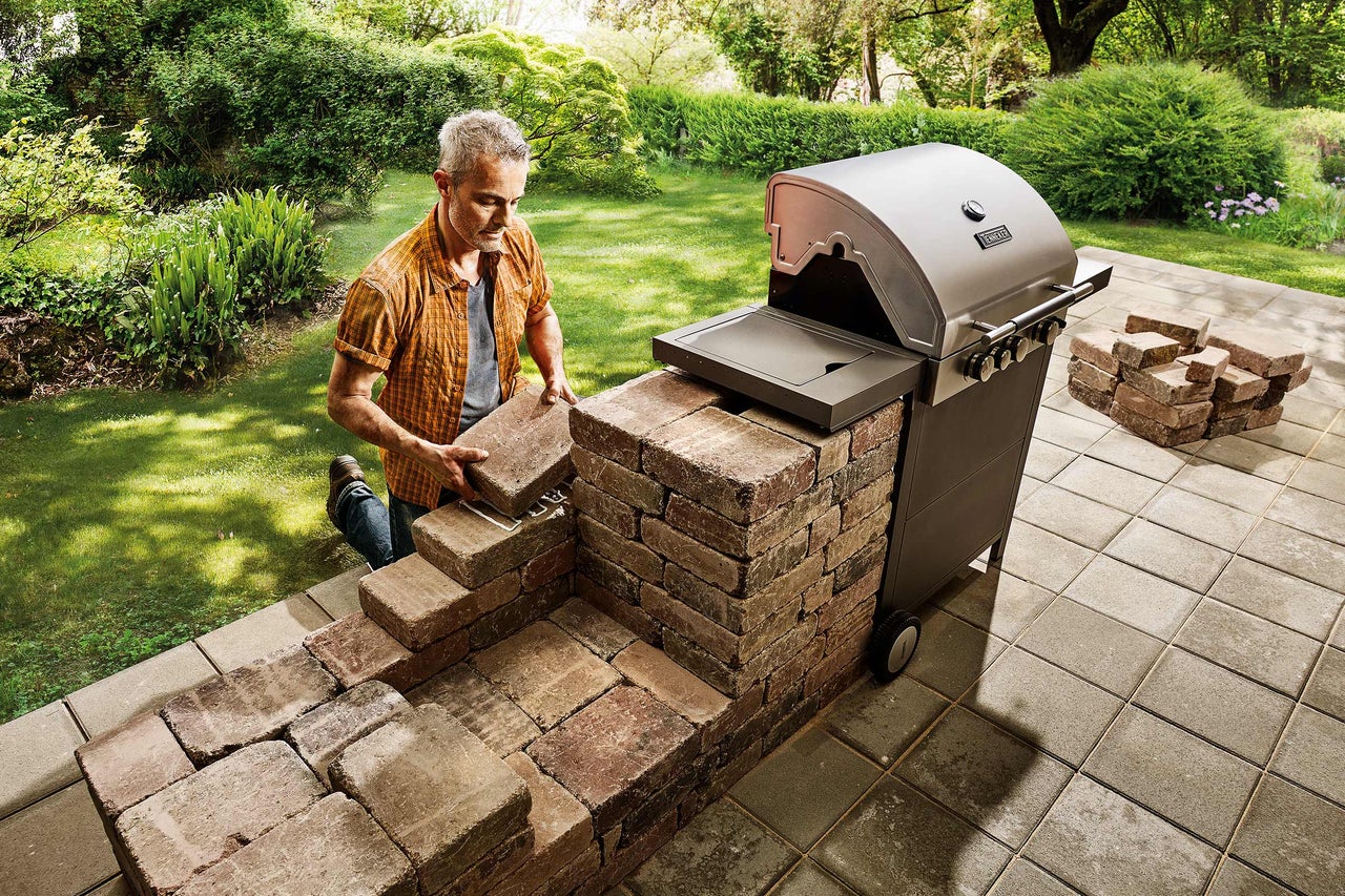 Un homme construit un barbecue en pierres à côté d'un barbecue à gaz dans le jardin.