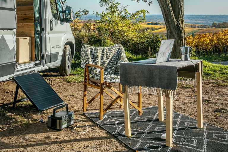 Scène en extérieur avec camping-car, panneau solaire, table, chaise et ordinateur portable dans un environnement naturel.