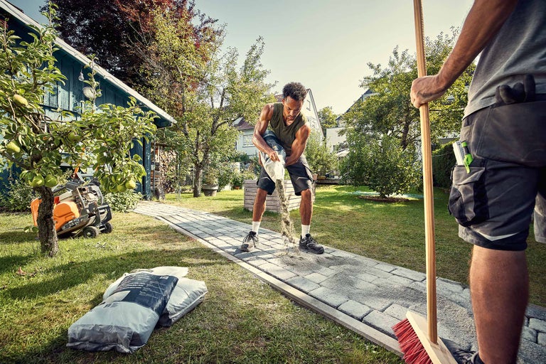 Landschaftsbild mit zwei Männern, die einen Pflasterweg mit Fugensand bearbeiten, mit einem Stampfer, Säcken mit Fugensand und einer Kehrmaschine