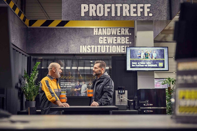Deux hommes se tiennent au Hornbach ProfiTreff avec des tasses Hornbach à une table.
