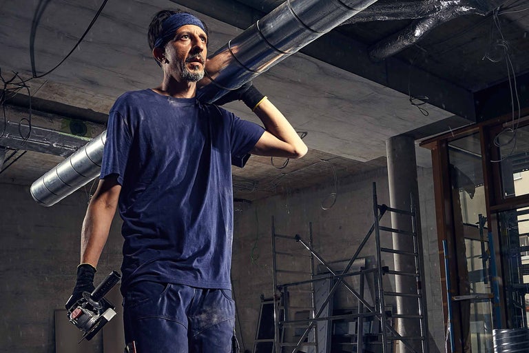 Un ouvrier porte un conduit de ventilation sur l'épaule, à l'arrière-plan une pièce inachevée avec un échafaudage.