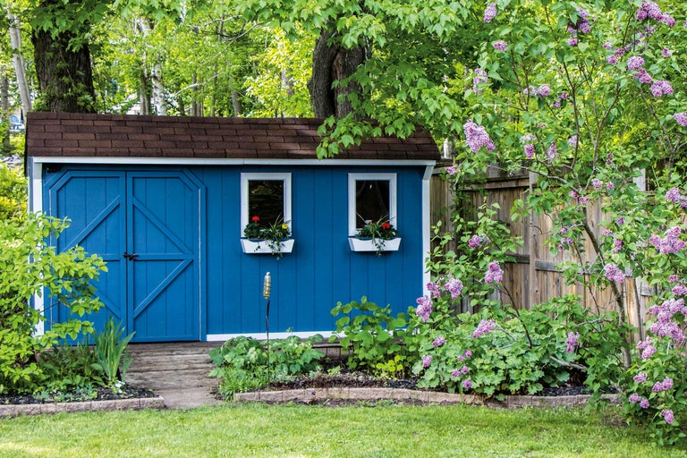 Gartenansicht mit blauem Geräteschuppen, Fenstern mit Blumenkästen und lila Blütensträuchern