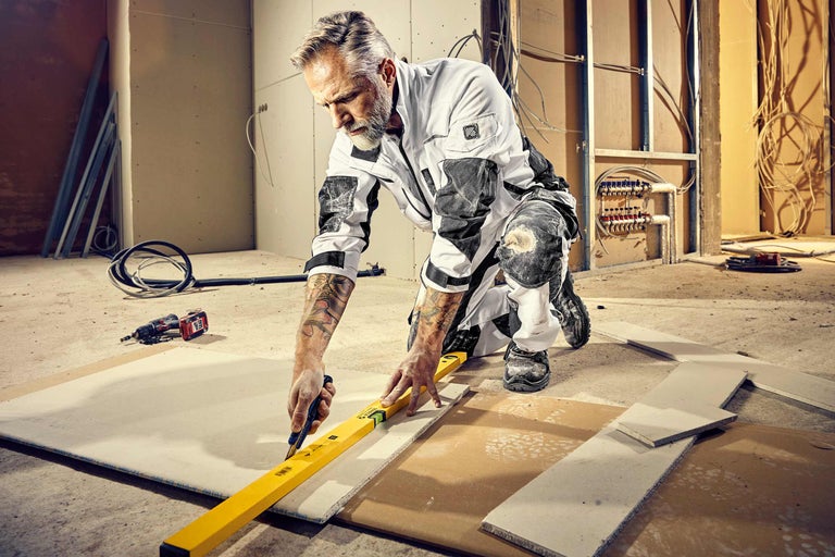 Un homme agenouillé dans une pièce en construction découpe une plaque de plâtre à l'aide d'un cutter le long d'un niveau à bulle. À l'arrière-plan, on aperçoit des tuyaux, des câbles et une perceuse.
