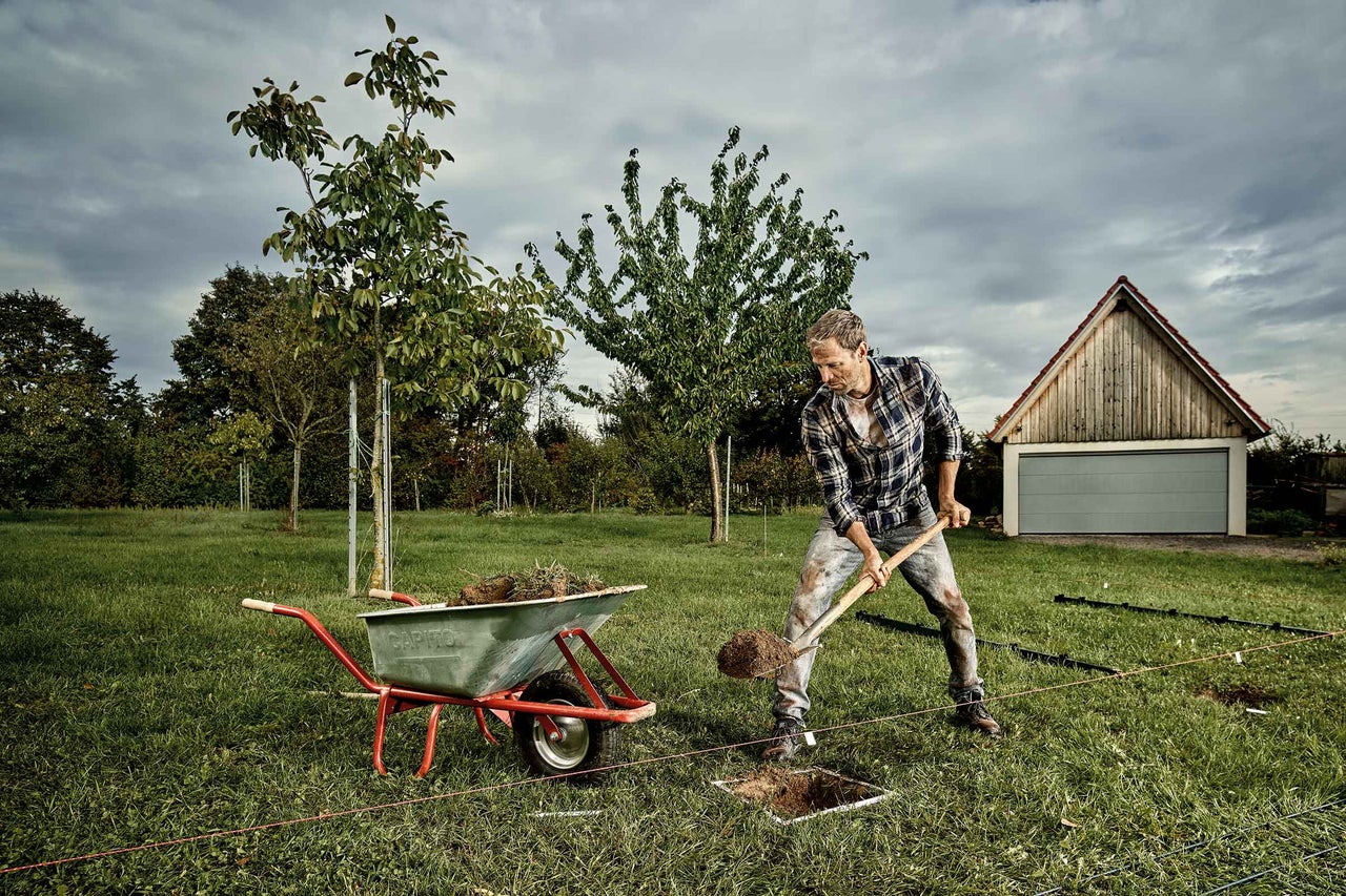 Un homme creuse un trou dans le jardin à côté d'une brouette avec une bêche.