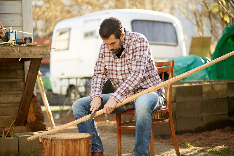 Un homme est assis sur une chaise et sculpte un bâton avec un couteau, une caravane est visible en arrière-plan.