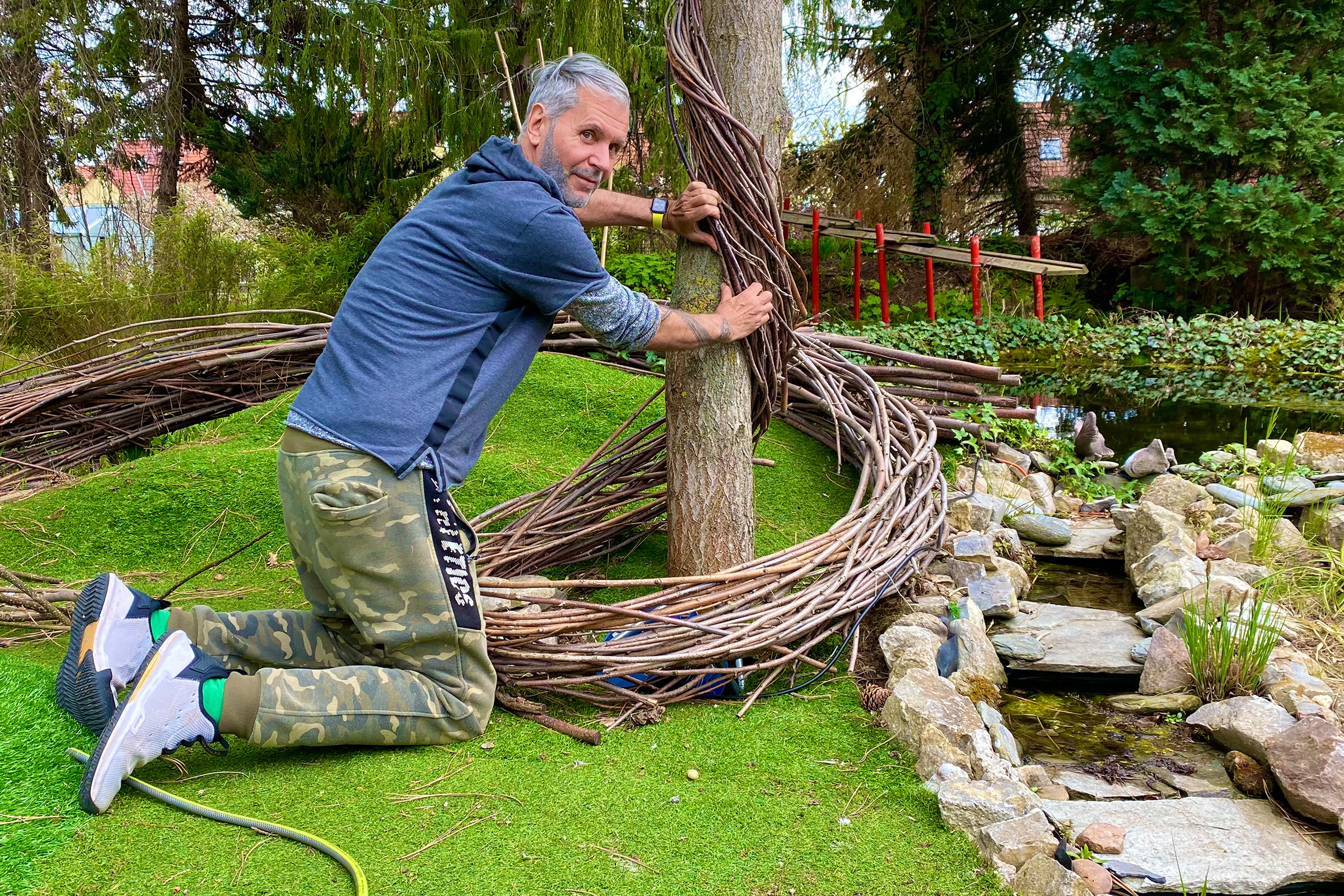 Mann befestigt Weidenkranz an Baum im Garten mit Teich und Bachlauf.