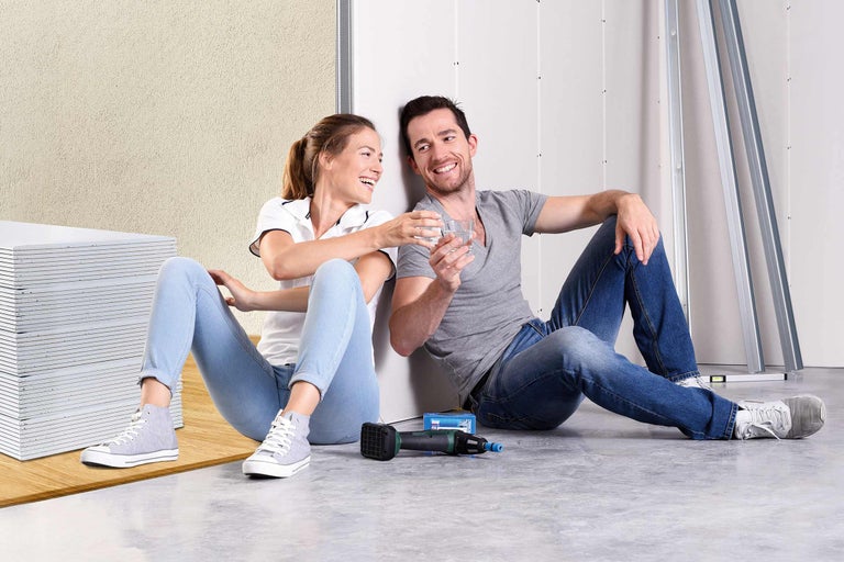 Ein Paar sitzt nach der Installation von Gipskartonplatten und Trockenbauprofilen mit einem Trockenbauschrauber auf dem Boden und hält Gläser mit Wasser.