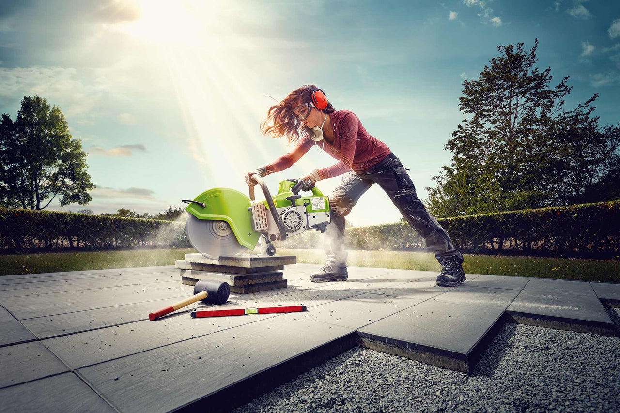 Une femme coupe des plaques de pierre avec une meuleuse d'angle à l'extérieur. Des équipements de protection individuelle sont utilisés.