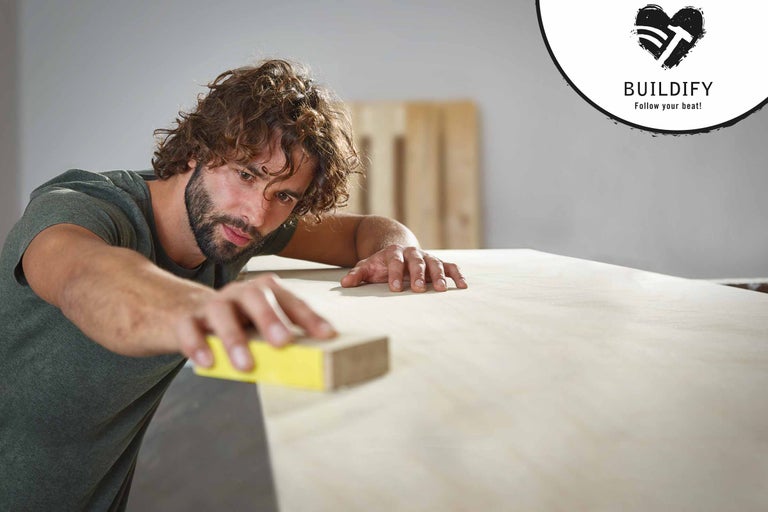 Un homme ponce du bois avec une cale à poncer. Logo Buildify.