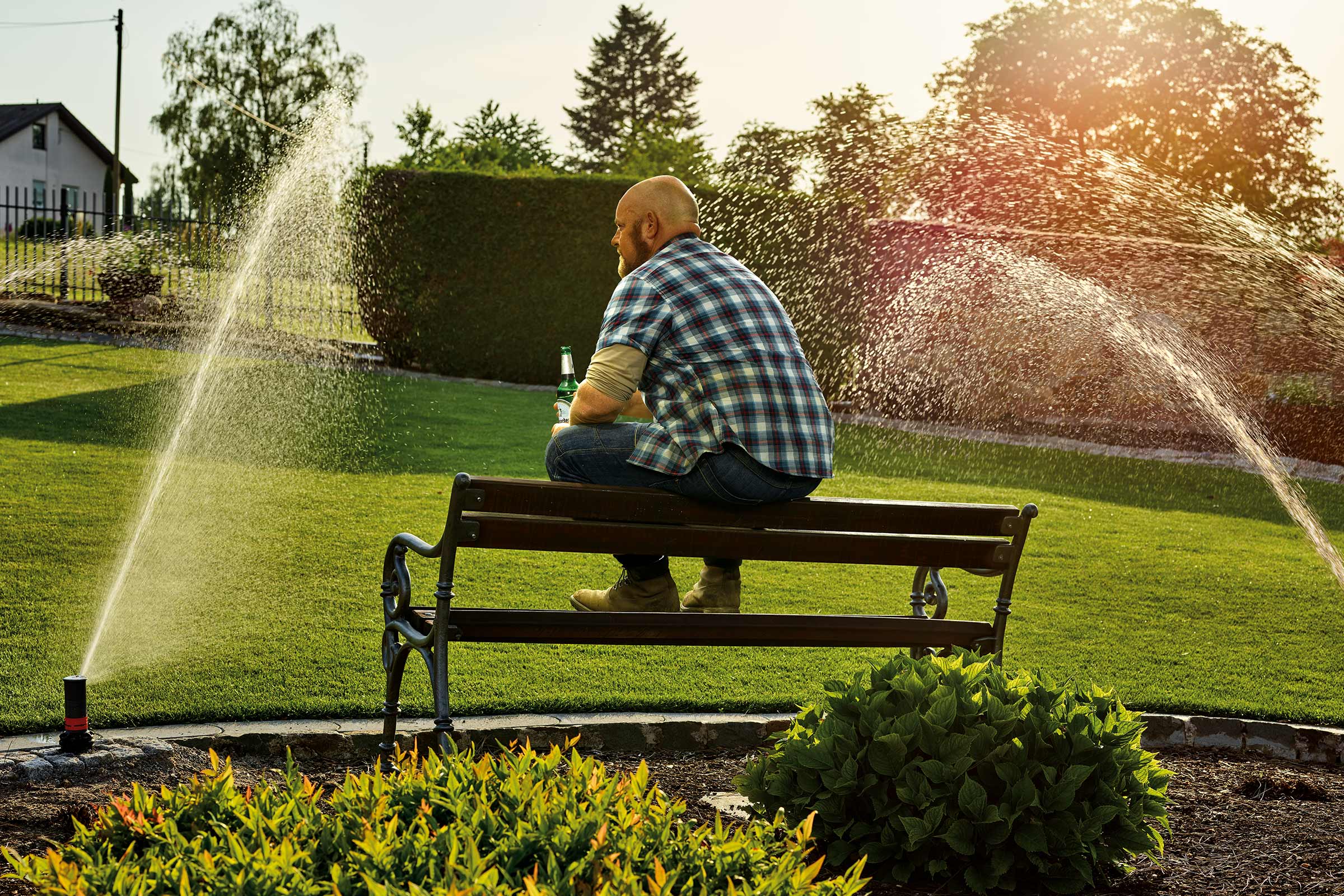 Un homme est assis sur un banc dans le jardin pendant qu'un arroseur arrose la pelouse.