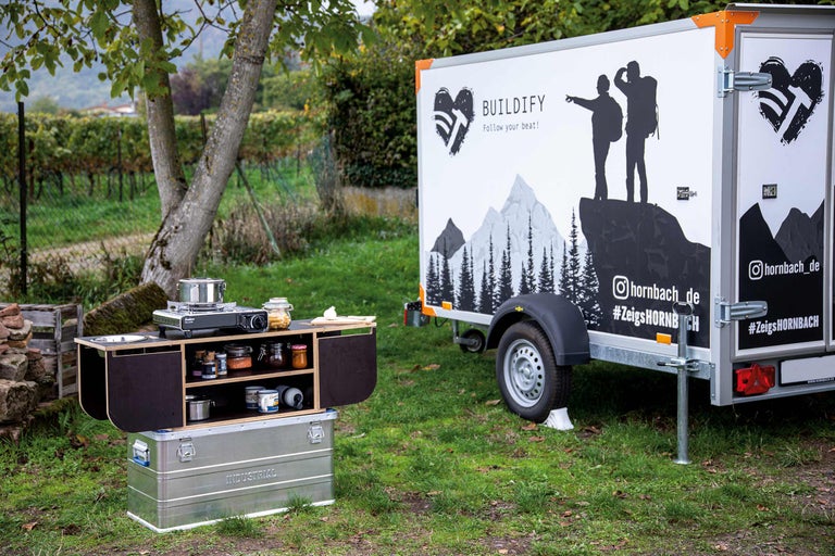 Cuisine de camping avec cuisinière, provisions et espace de rangement sur un terrain avec une remorque en arrière-plan.