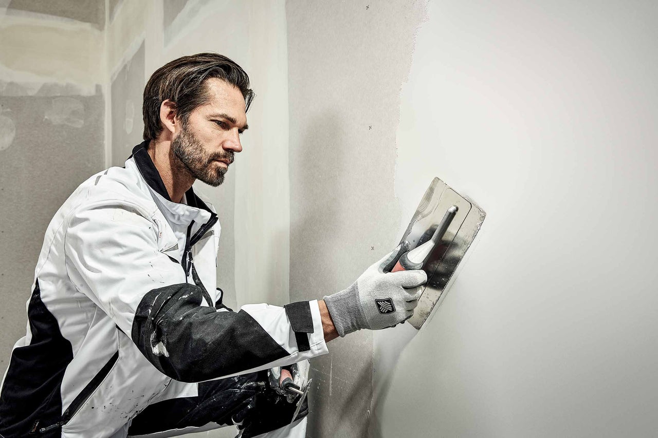 Ein Mann glättet eine Wand mit einer Glättkelle. Die Wand ist teilweise verputzt.