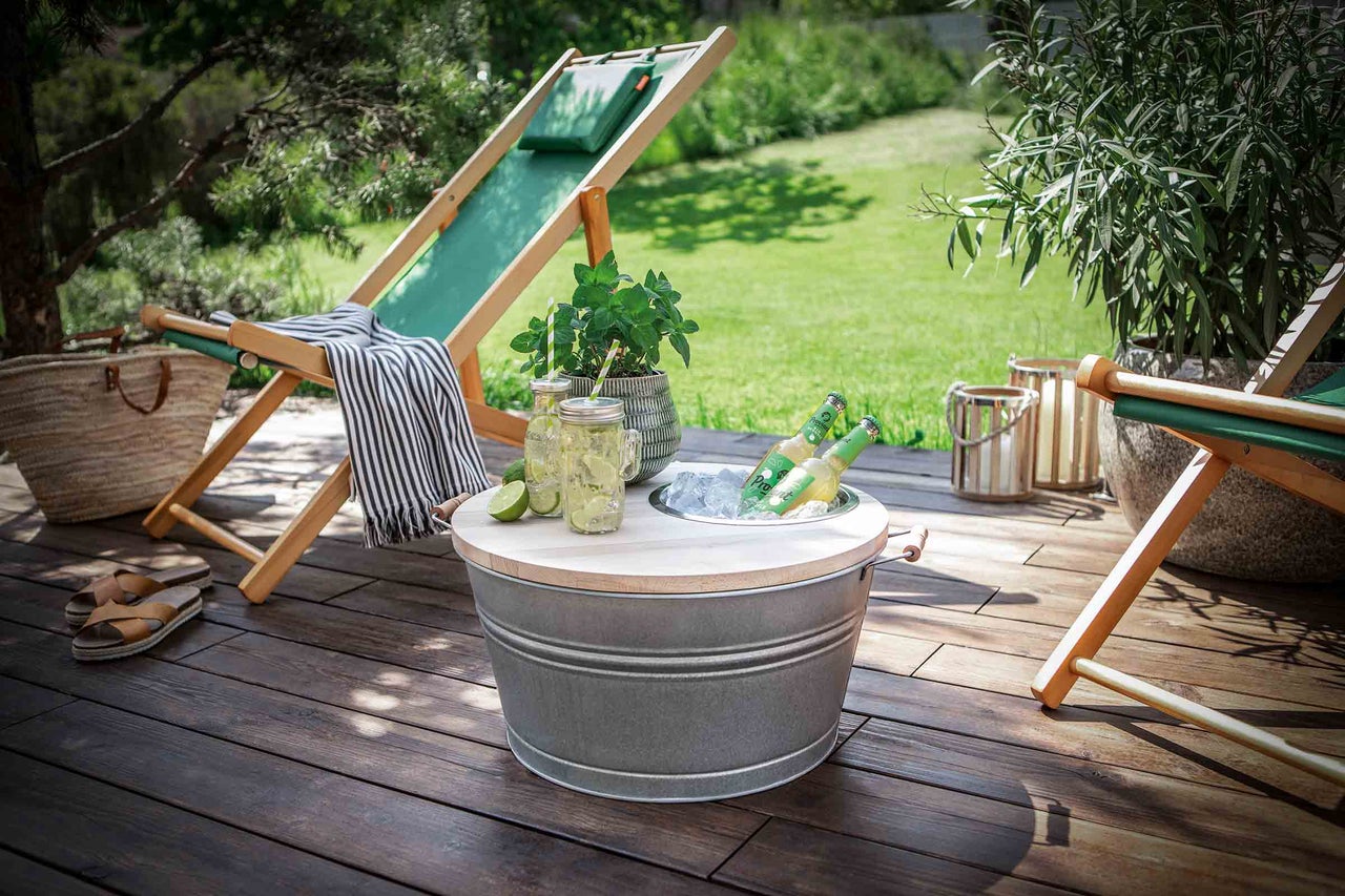 Scène de jardin estivale avec chaises longues, rafraîchisseur de boissons avec glace et boissons, verres de limonade, plantes et décoration sur une terrasse en bois