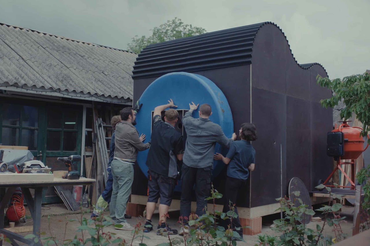 Un groupe de personnes monte un élément circulaire sur le mur extérieur d'une petite maison en bois moderne avec un toit ondulé dans un jardin.