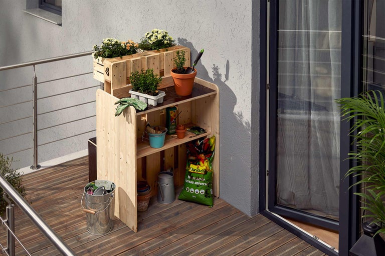 Gartenszene mit einem Pflanztisch aus Holz, Blumenkästen, Pflanzen und Gartengeräten auf einem Balkon
