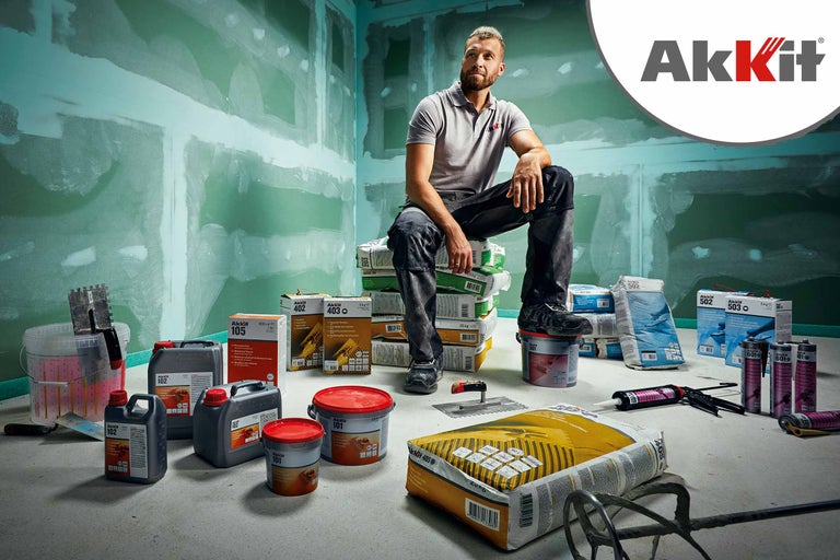 Présentation de divers produits Akkit, notamment des mastics, des enduits et des adhésifs, devant un chantier avec des murs plâtrés. Un homme est assis sur une pile de sacs Akkit. Logo Akkit.
