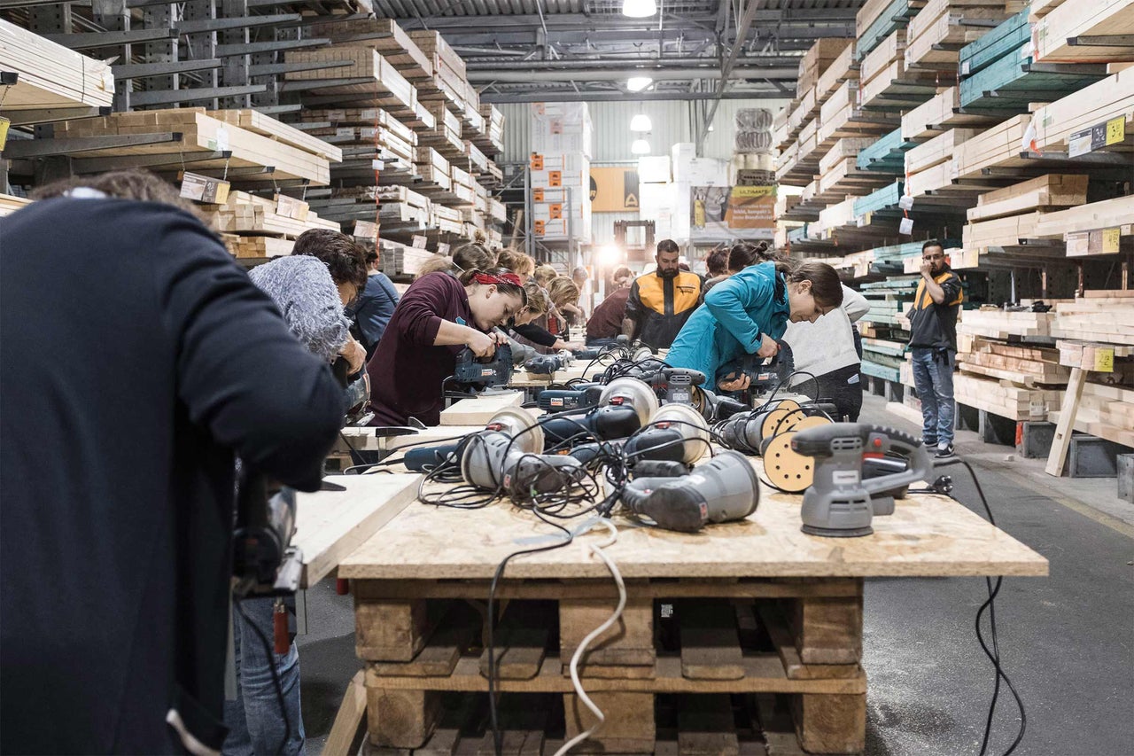 In einer Hornbach Filiale sägen und schleifen mehrere Personen Holz an einem Tisch mit Elektrowerkzeugen vor Holzregalen.
