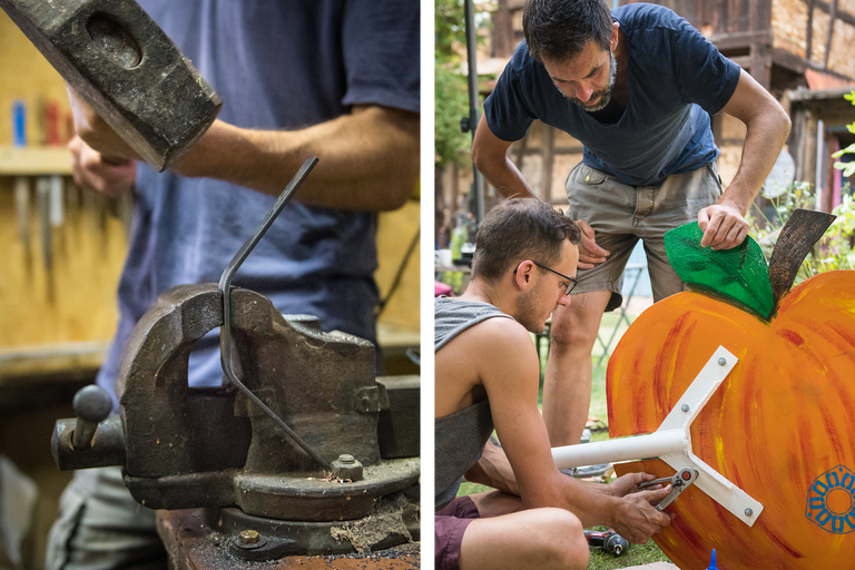 Zwei Männer arbeiten an einem Metallstück mit einem Hammer und einer Rohrzange sowie an einer großen, bemalten Holzfrucht