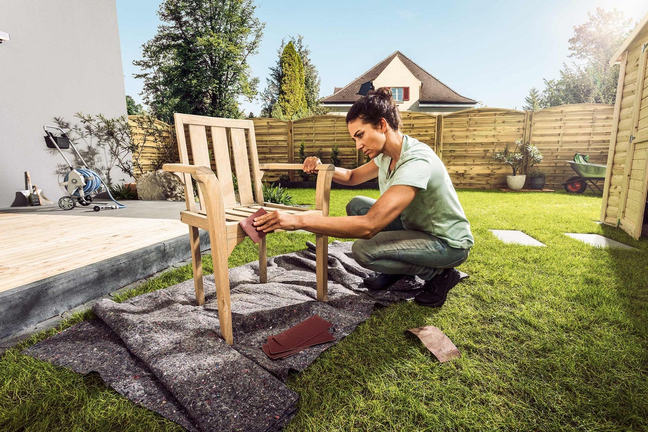 Une femme ponce une chaise en bois dans le jardin avec du papier de verre.