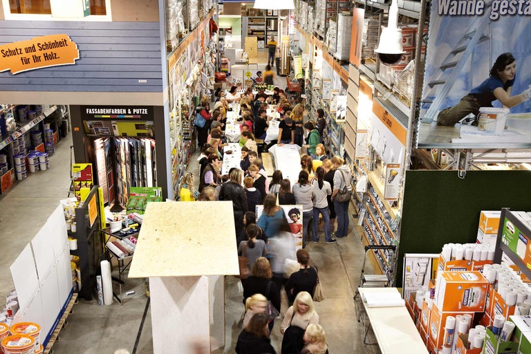 Intérieur d'un magasin de bricolage Hornbach avec des clients assis à des tables, entourés de produits tels que peintures, enduits et papiers peints.