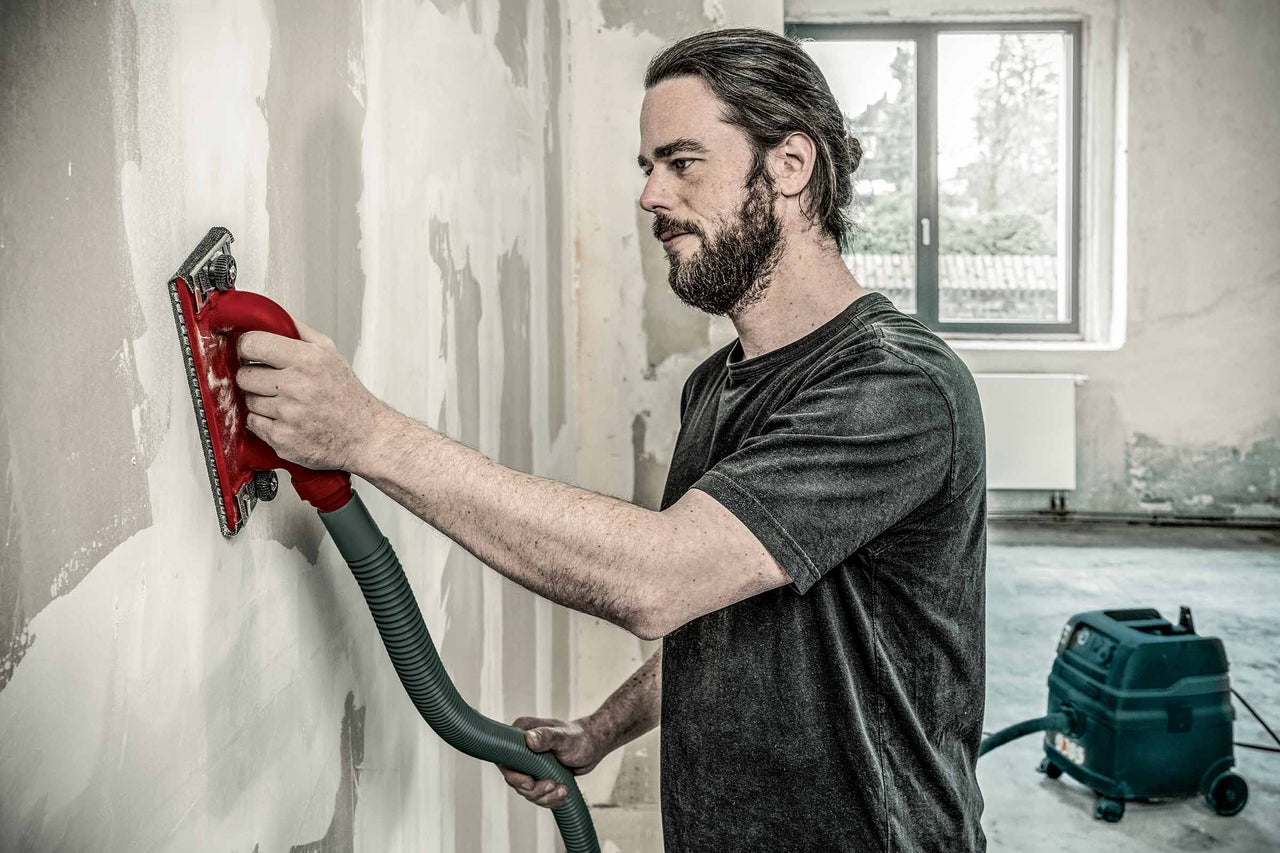 Mann schleift Wand mit einem Trockenbauschleifer, angeschlossen an einen Staubsauger