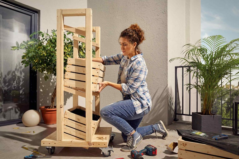 Une femme assemble un potager surélevé mobile en bois sur un balcon avec des outils et des plantes.
