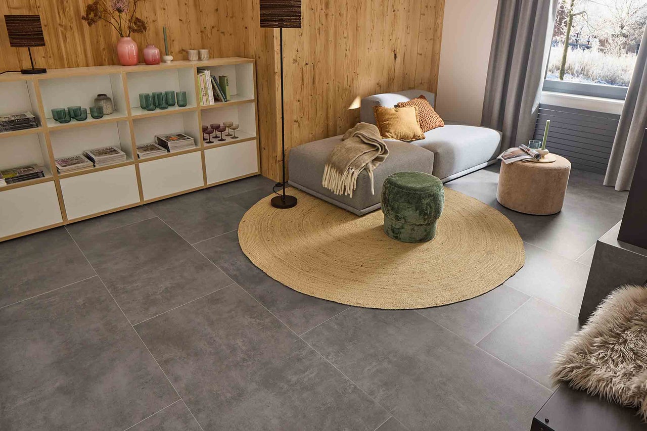 Salon avec carrelage gris, canapé, tapis, tabouret et étagère.