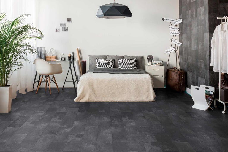 Chambre à coucher avec lit, bureau et revêtement de sol foncé aspect pierre.