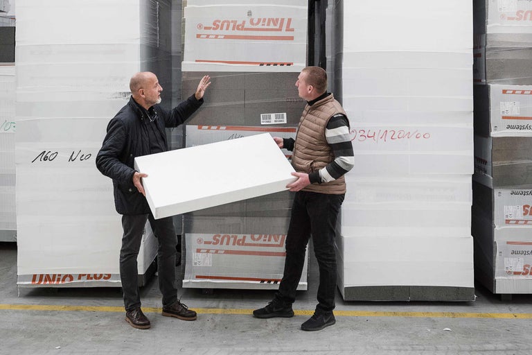 Zwei Personen hantieren mit einer Wärmedämmplatte aus Polystyrol vor einem Stapel verpackter Dämmplatten.