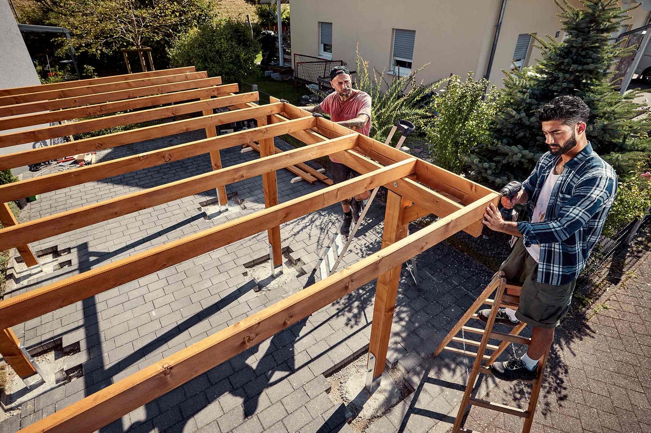 Deux artisans assemblent une structure en bois pour une toiture de terrasse avec une visseuse sans fil sur une terrasse pavée dans le jardin.