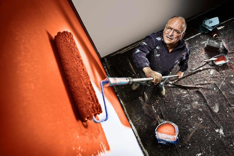 Un homme peint un mur avec un rouleau à peinture, un pot de peinture et une bâche sont également visibles sur l'image.