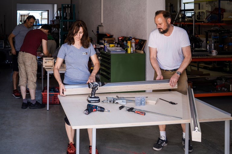 Personen arbeiten mit Bohrmaschinen, Schrauben, Zwingen und Holz auf einer Werkbank.