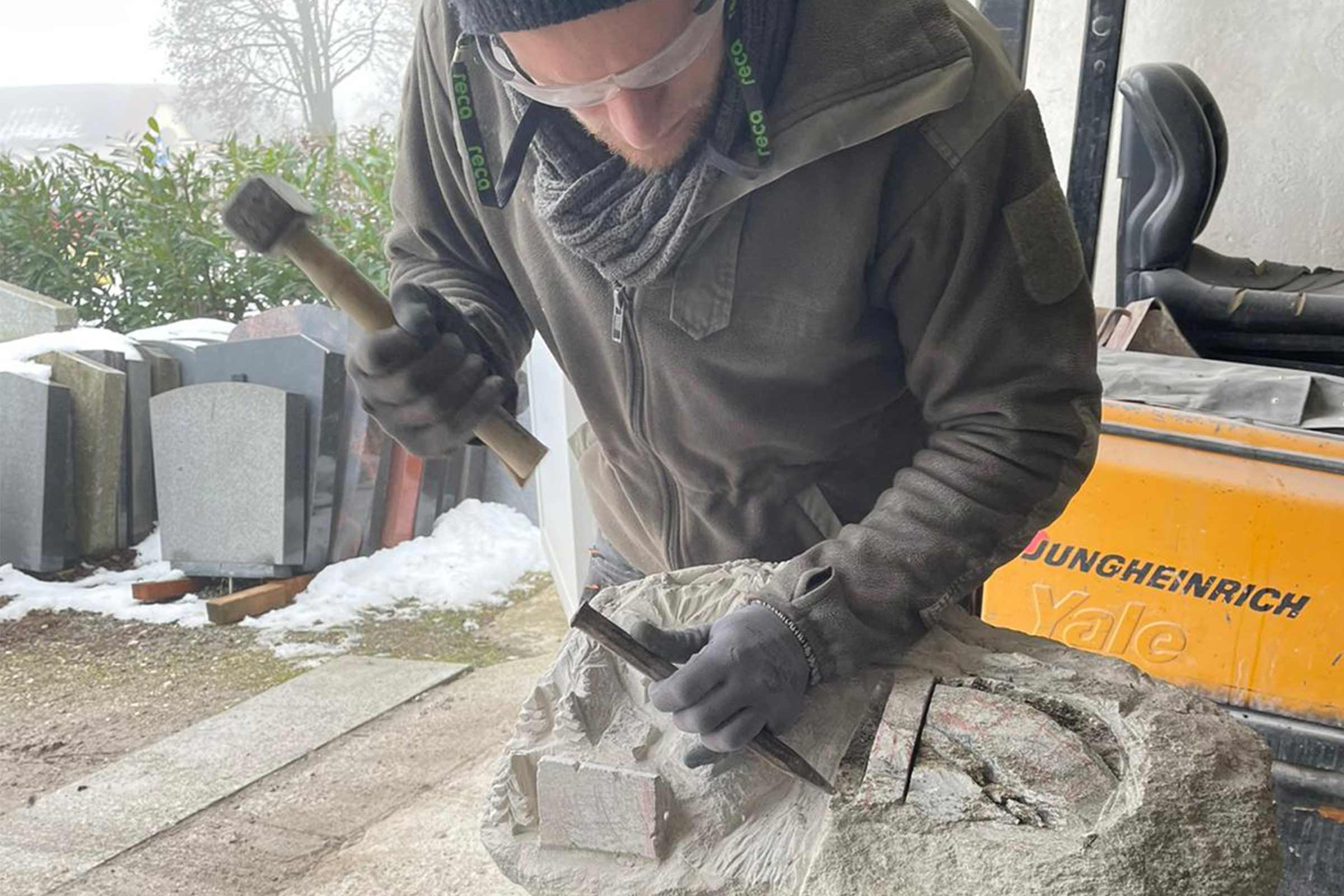 Ein Mann bearbeitet einen Stein mit Hammer und Meißel, im Hintergrund Grabsteine.