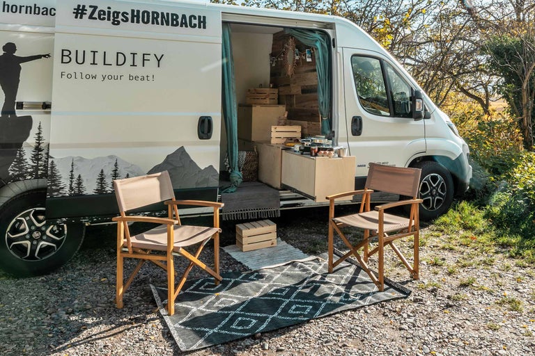 Szene mit einem weißen Van, vor dem zwei Holzstühle auf einem gemusterten Teppich stehen