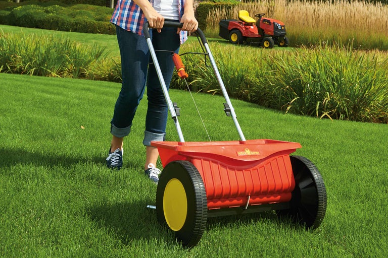 Une femme utilise un épandeur sur une pelouse. Un tracteur de jardin est visible à l'arrière-plan.