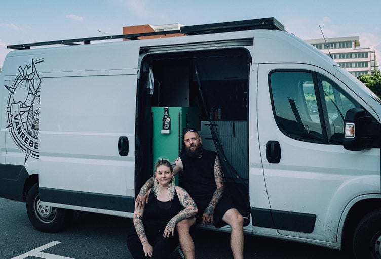 Ein Paar sitzt vor einem Transporter mit offenstehender Tür. Auf der Seite des Fahrzeugs befindet sich ein Logo.