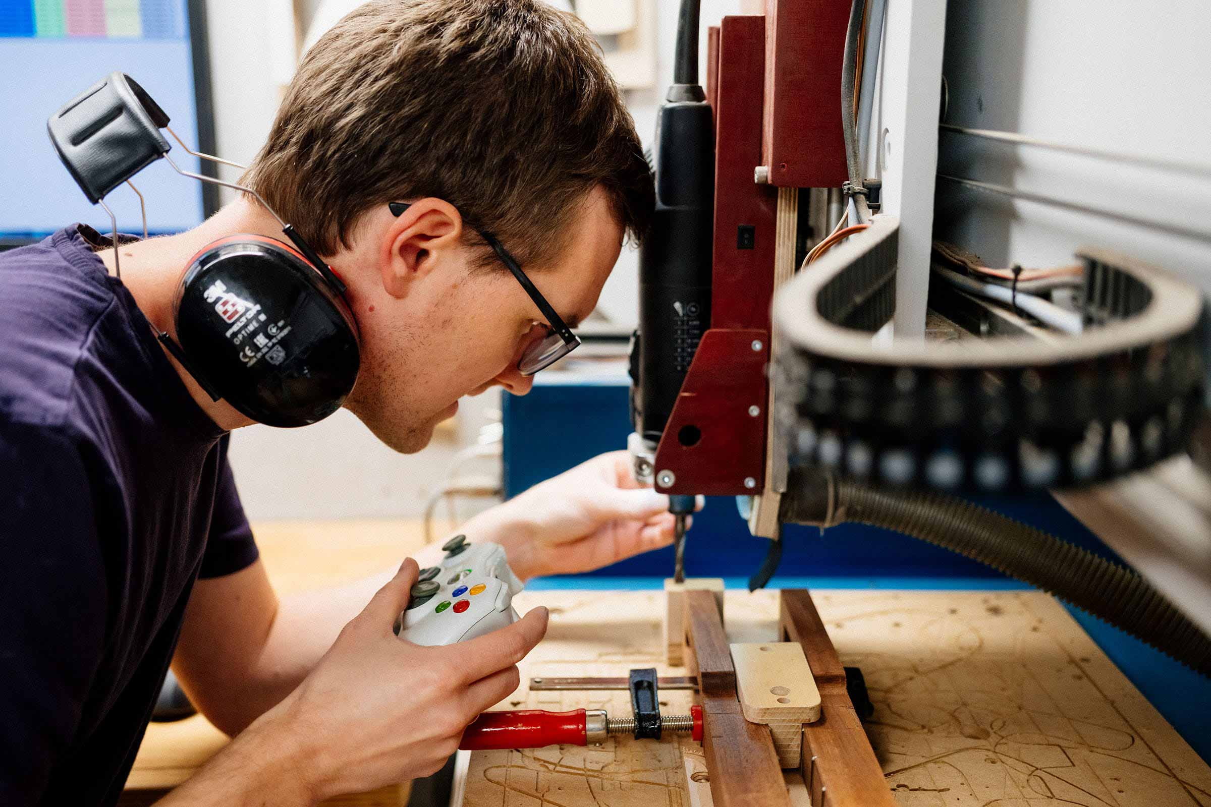 Ein Mann mit Kopfhörern und Brille bedient eine CNC-Fräse mit einem Gamecontroller.