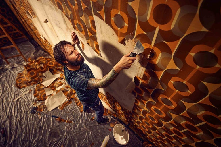 Un homme retire du vieux papier peint d'un mur à l'aide d'un grattoir, tandis qu'une bâche recouvre le sol.