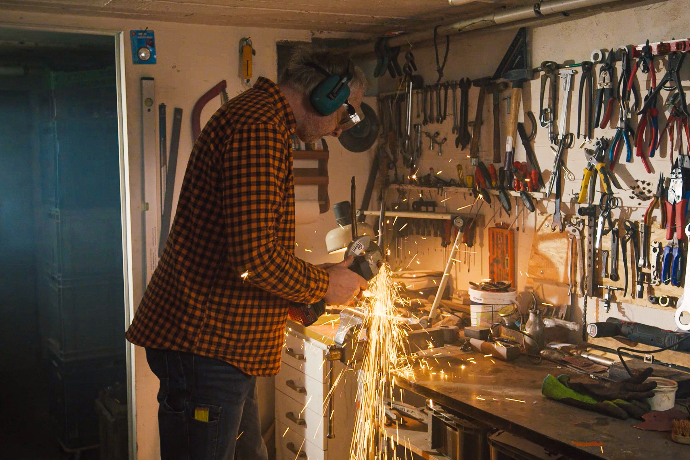 Mann mit Gehörschutz arbeitet mit einem Winkelschleifer in einer Werkstatt, bei der Funken fliegen und Werkzeuge an der Wand hängen.