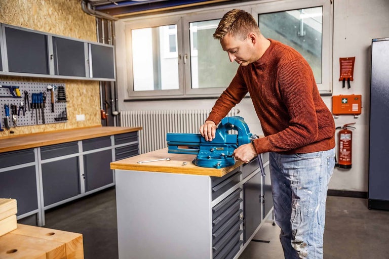 Mann montiert blauen Schraubstock auf Werkbank in einer hellen, organisierten Werkstatt mit Werkzeugwand, Hängeschränken und Holzoberflächen.