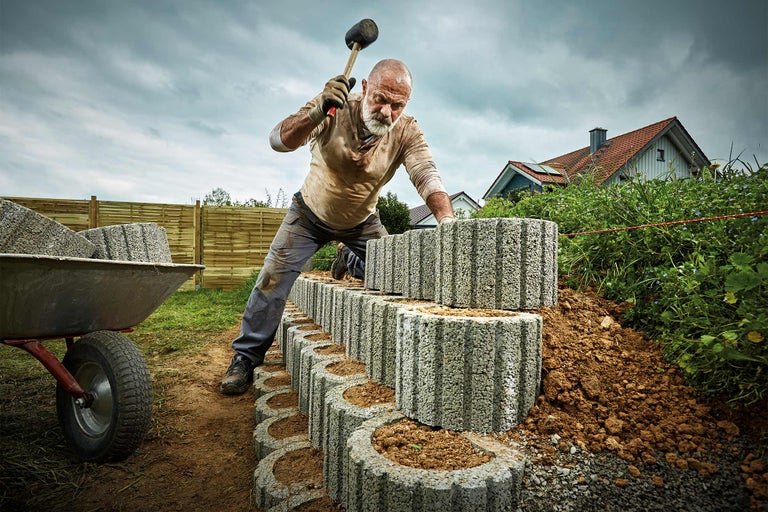 Un homme construit un mur de jardin en pierres de béton avec un maillet en caoutchouc, une brouette en arrière-plan.