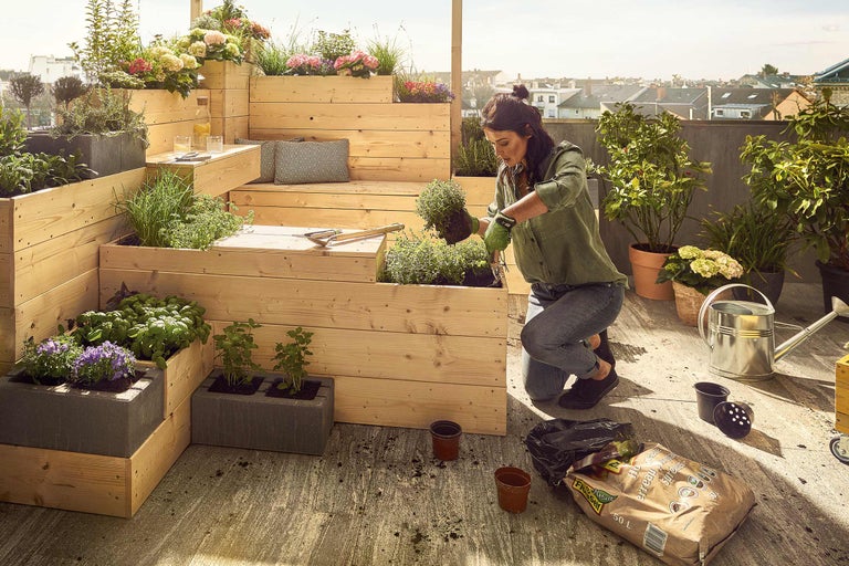 Frau bepflanzt Hochbeet auf einer Dachterrasse mit verschiedenen Kräutern und Blumen.