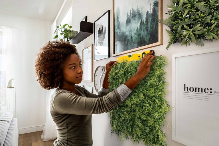 Une femme installe un mur végétal vert à l'aide d'un niveau à bulle sur un mur, entourée de décorations. Ambiance d'espace lumineuse et organisée.