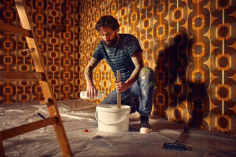 Un homme mélange de la peinture dans un seau pour des travaux de rénovation dans une pièce avec du papier peint rétro.