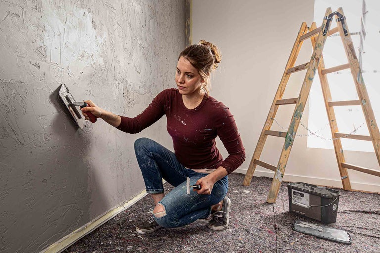 Frau trägt Betonspachtel mit einer Glättkelle auf eine Wand auf, neben einer Leiter.
