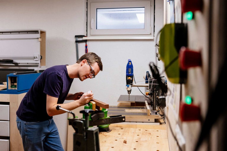 Ein Mann arbeitet an einem Holzstück mit einer Werkbank, einer Schraubzwinge und einer Standbohrmaschine in einer Werkstatt.