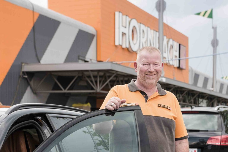 Un collaborateur Hornbach se tient devant une voiture, devant le magasin de bricolage Hornbach.