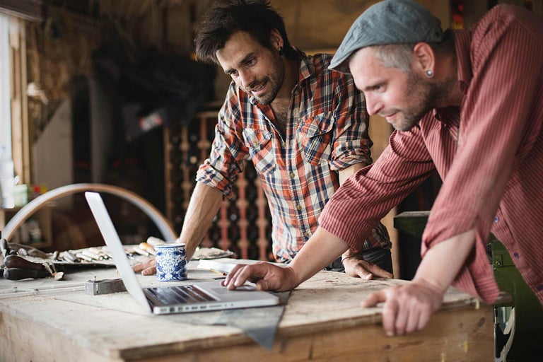 Deux artisans travaillent sur un ordinateur portable dans un atelier. Des outils et des matériaux de construction sont visibles en arrière-plan.