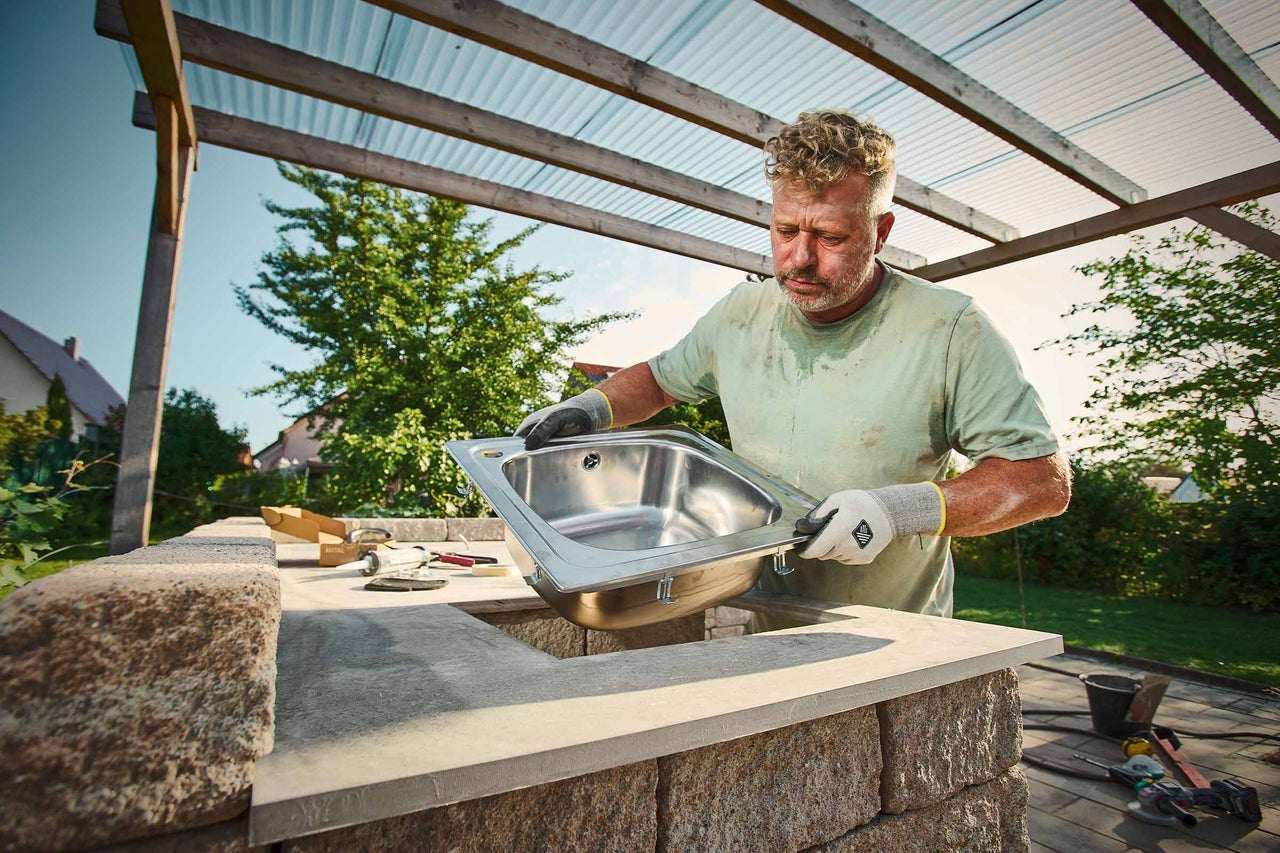 Un homme installe un évier à encastrer en acier inoxydable dans un plan de travail en pierre d'une cuisine d'été.